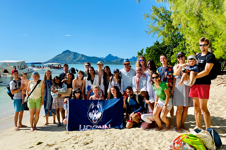 A group of students on a sunny beach holding a UConn flag.